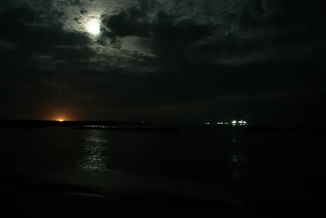 夜のふるさと海岸。ほぼ真っ暗で船や月の光だけが見える。オレンジの光はおそらく地上の外灯。